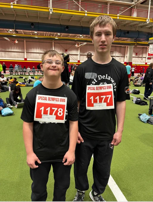 Two young athletes wearing black shirts and Special Olympics Iowa bibs 1177 and 1178 standing on an indoor track field.
