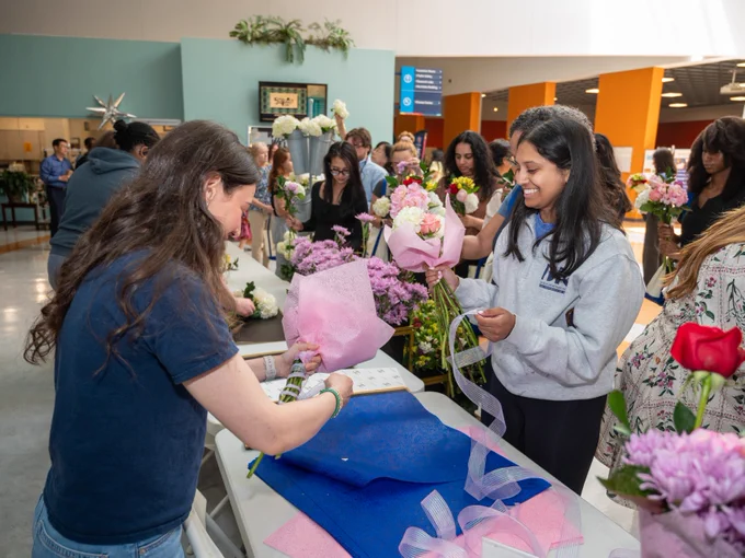 A group of people are engaged in assembling flower bouquets at a table. Various flowers and decorative materials are laid out. The setting appears to be indoors, possibly at an organized event or workshop.