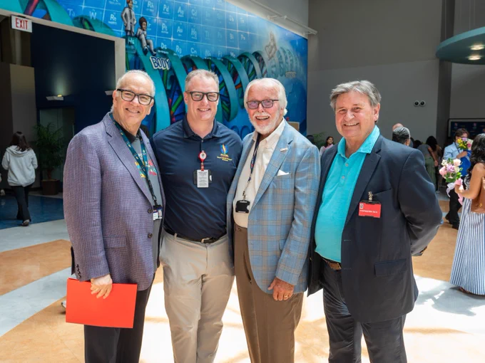 A group of four people are standing together indoors, smiling at the camera. They are dressed in business casual attire. Behind them, a colorful mural featuring various scenes and designs is visible on the wall. The setting appears to be a formal or professional event.