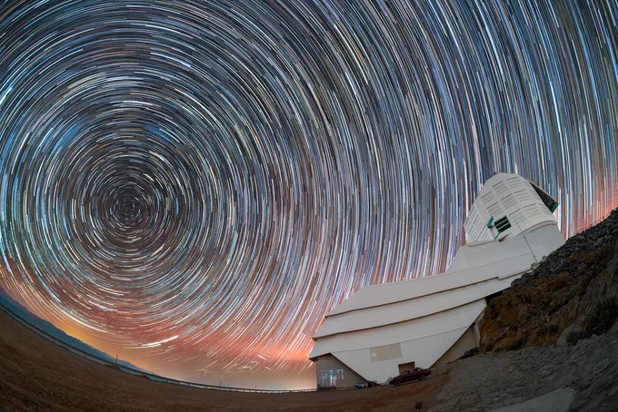 Concentric star trails curve across a night sky above a large boot-shaped white observatory on a rocky desert slope. A warm twilight glow lights the horizon.