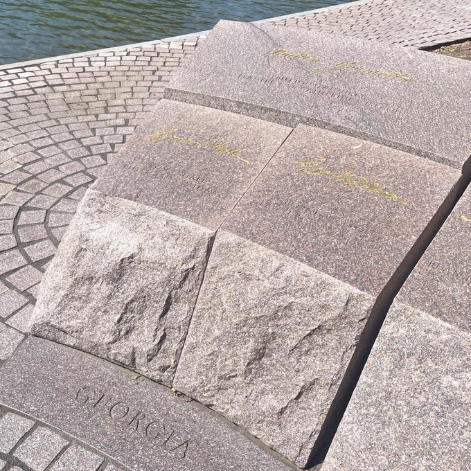 Three reddish gray stone blocks with names etched into the tops and gold signatures on them and the word "Georgia" written at the base stand as a section of a low memorial wall.