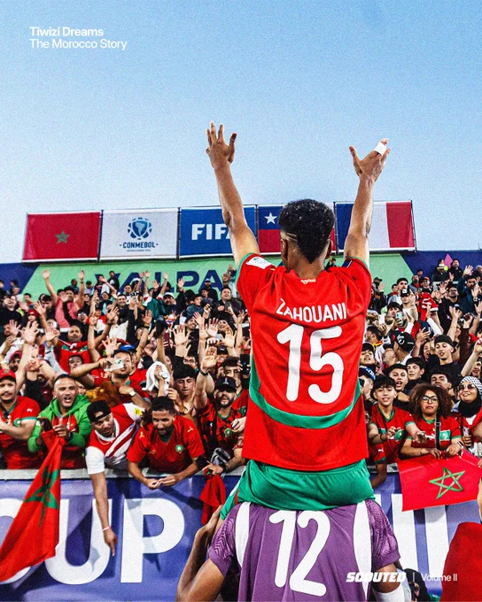 Photo of Faouad Zahoiani being lifted on a teammate's shoulders, celebrating a victory at the 2025 FIFA U-20 World Cup. He's wearing a vibrant red shirt with 'ZAHOUANI 15' on the back and gesturing with both arms up in front of Moroccan fans. Above him are the flags or Morocco and France set against a hazy blue sky.
