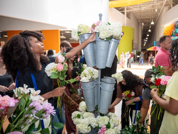 People are gathered around a stand holding flower bouquets, selecting various flowers like hydrangeas. The scene takes place indoors with colorful walls in the background.