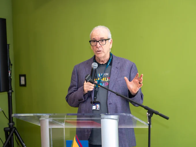 A person is speaking into a microphone at a clear podium against a green background. They are wearing glasses, a T-shirt with a logo, and a blazer.