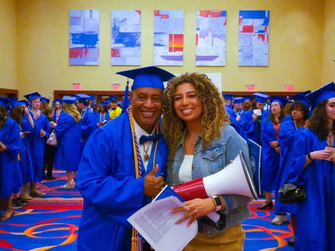 Graduates in blue gowns and caps celebrate in a colorful room. One individual gives a thumbs-up while holding a diploma. Another person beside them holds a megaphone. 
