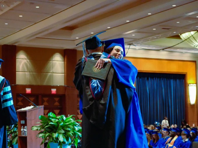 A graduate in a blue cap and gown receives a hug while on stage, holding a diploma. The event takes place in a large auditorium with seated attendees wearing similar attire.