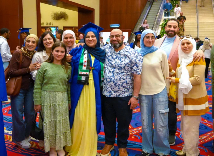 A group of people are gathered together in a celebratory setting. One person is wearing a blue graduation cap and gown, with a colorful sash. The atmosphere appears joyful.