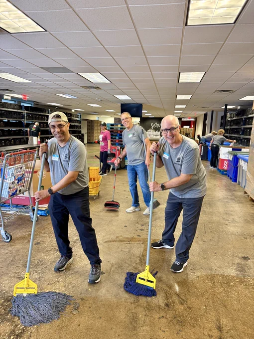 A group of three smiling individuals, wearing matching gray shirts, is seen cleaning the floor with mops in a large indoor space. Shelves with various food items are visible in the background alongside a shopping cart and cleaning buckets. More volunteers can be seen working further back in the room.