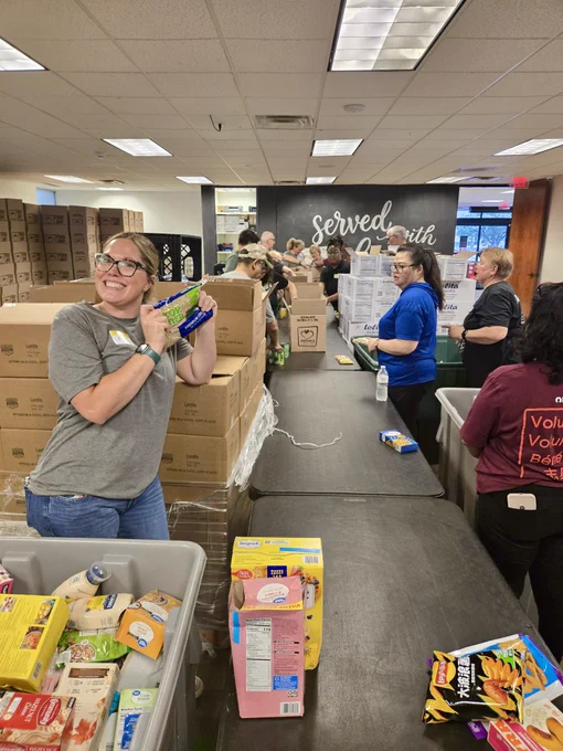 Volunteers are packing food supplies in a large room with numerous cardboard boxes. A woman in the foreground is holding a box of macaroni and smiling. In the background, more people are organizing items on a long table. The wall displays the words "Served with Heart.