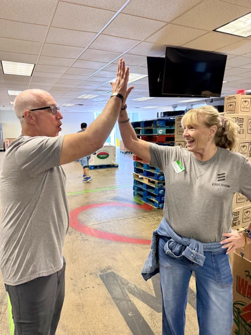 A man and a woman, both wearing gray Ericsson shirts, high-five each other in a food pantry filled with pallets and boxes. There is a TV hung on the ceiling on the right side. 