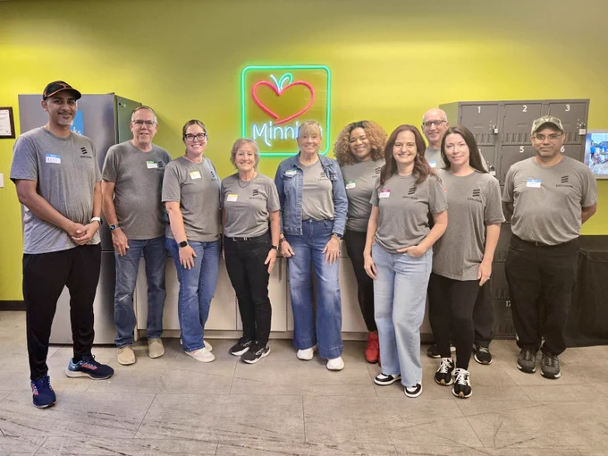 A group of 10 people standing and smiling in front of a bright yellow wall with a green neon sign featuring a red heart and the word "Minnie's" in white. They are wearing matching gray T-shirts with a black Ericsson logo on the right side and white name tags on the left. 