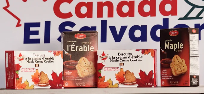 Maple cookie and cream products displayed in front of a backdrop reading “Canada El Salvador,” with packaging featuring maple leaves and syrup bottles.