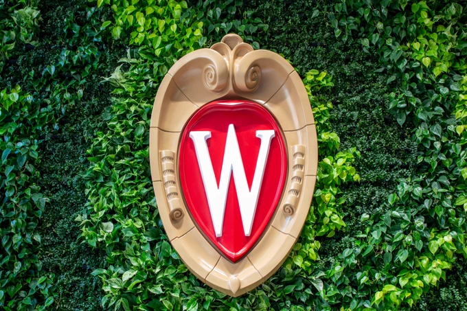A large UW-Madison crest is mounted on a green wall made of ferns and pothos. The original can be found on the ground floor of Morgridge Hall.