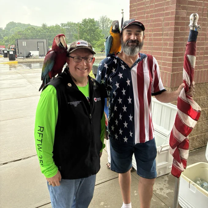 A Red Cross volunteer stands next to another person wearing a patriotic shirt. They both have a parrot on their shoulder.