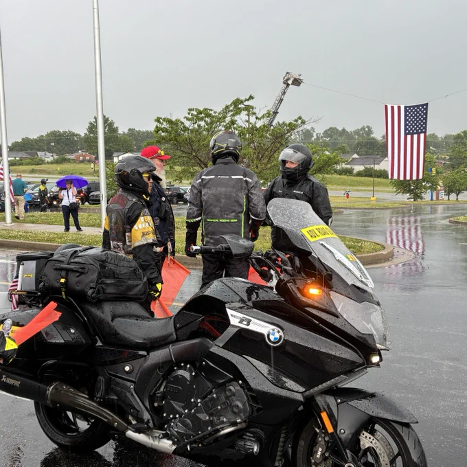 A shot of a motorcycle and three riders in helmets. In the distance, a large American Flag is hung between two cranes above a paved path.