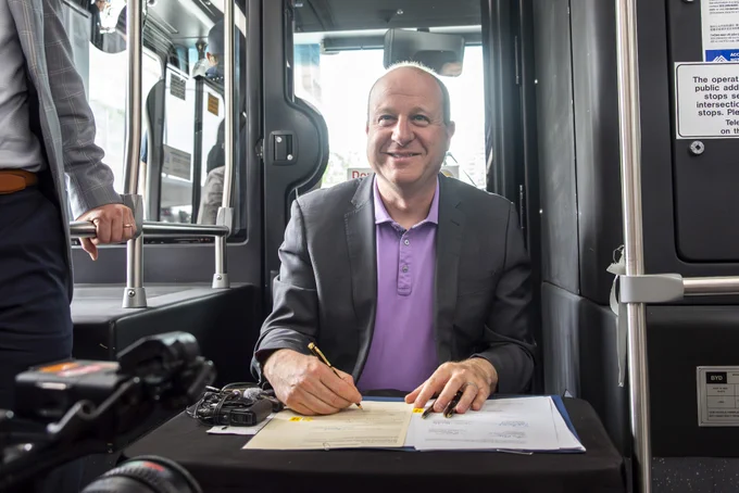 Man in a blazer and purple shirt sitting at a table inside a bus, writing on documents with a pen.