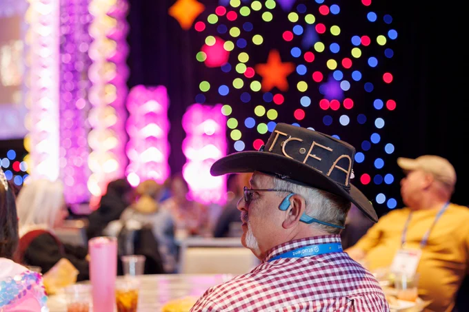 Photo of a person's side profile as they sit at a table. They have a hat on with the word "TCEA" decorating the side of the hat. The background is blurry with colorful lights, other tables, and people. 