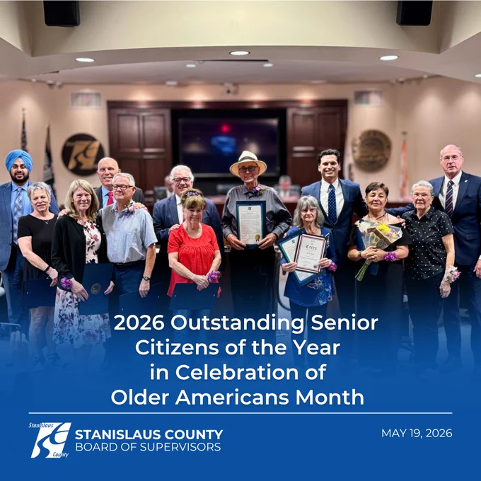 Group photo of the 2026 Outstanding Senior Citizens of the Year standing in a county board chamber during a recognition ceremony for Older Americans Month. Several seniors hold framed certificates and flowers while officials and community members stand alongside them. A blue graphic overlay at the bottom displays the text “2026 Outstanding Senior Citizens of the Year in Celebration of Older Americans Month,” along with the Stanislaus County Board of Supervisors logo and the date May 19, 2026.