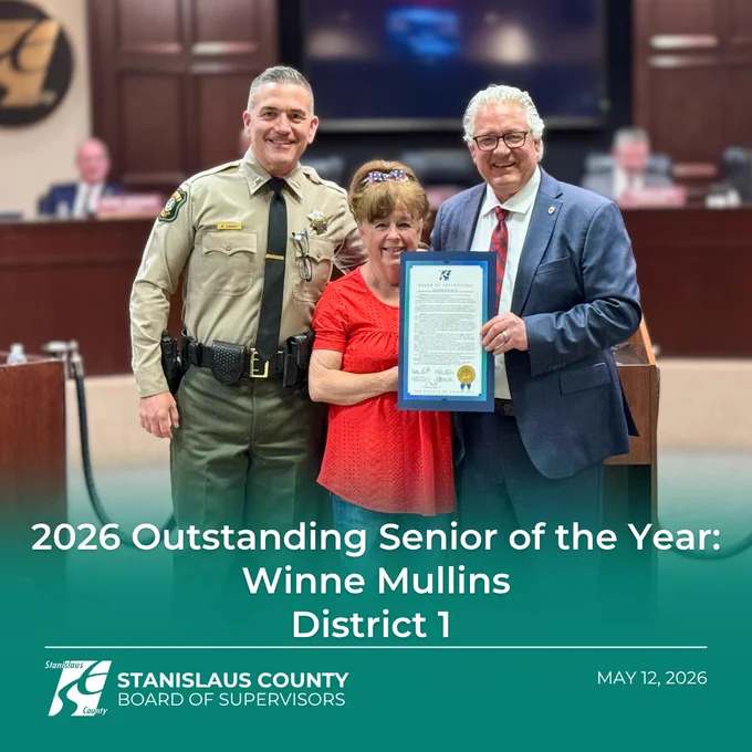 A woman in a red blouse stands between a uniformed sheriff’s officer and a county official during an award ceremony in a government board chamber. The official holds a framed certificate while all three smile for the photo. Text overlay reads “2026 Outstanding Senior of the Year: Winne Mullins, District 1,” with the Stanislaus County Board of Supervisors logo and the date May 12, 2026 at the bottom.