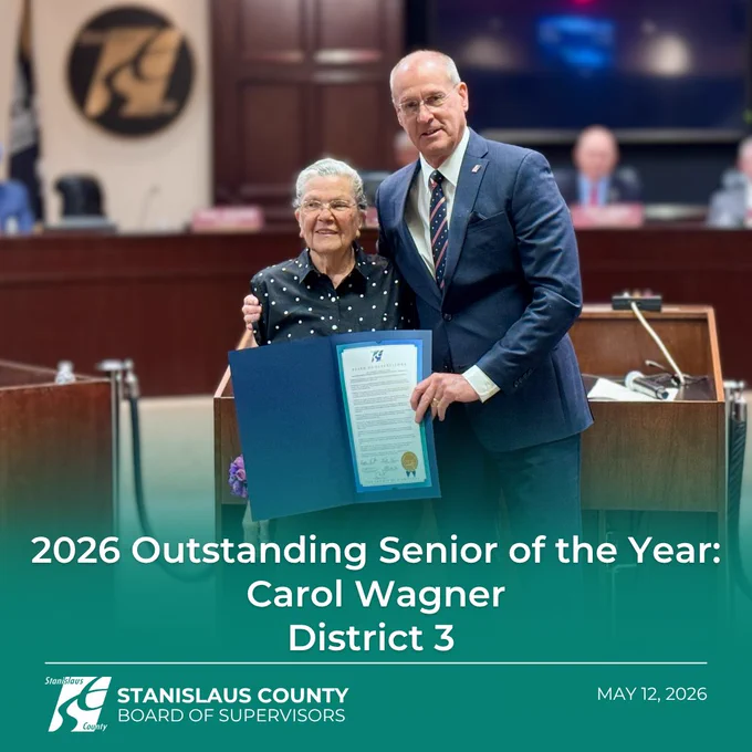 An older woman and a county official stand together in a government board chamber during an award presentation ceremony. The woman holds an open blue certificate folder while the official stands beside her with his arm around her shoulder. Text overlay reads “2026 Outstanding Senior of the Year: Carol Wagner, District 3,” with the Stanislaus County Board of Supervisors logo and the date May 12, 2026 at the bottom.

