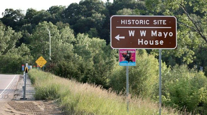 A roadside scene features a brown road sign indicating a historic site, the "W. W. Mayo House," with an arrow pointing left. An additional sign reads "Scenic Byway - Minnesota River Valley" with an image of an eagle. Trees and tall grass stretch along the roadside.