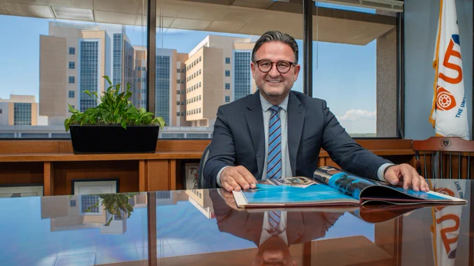 Dr. Jochen Resier sits at a shiny desk and smiles as he places his hands on an open book.  The background is the Jennie Sealy Hospital,