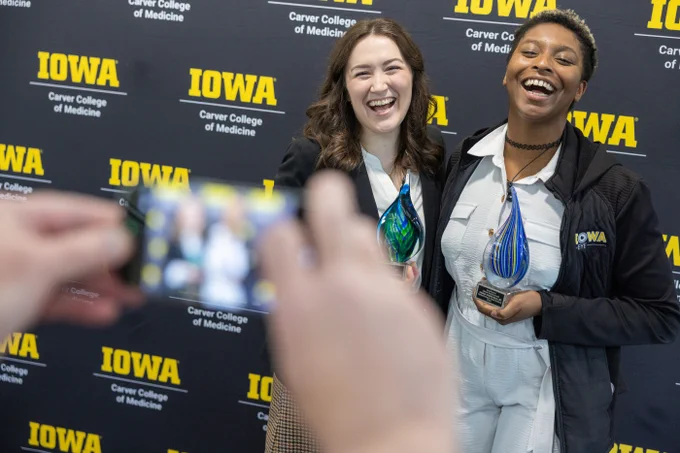 Two people stand smiling in front of a backdrop reading "Iowa Carver College of Medicine." They hold colorful glass awards. A hand is visible, capturing the scene with a smartphone.