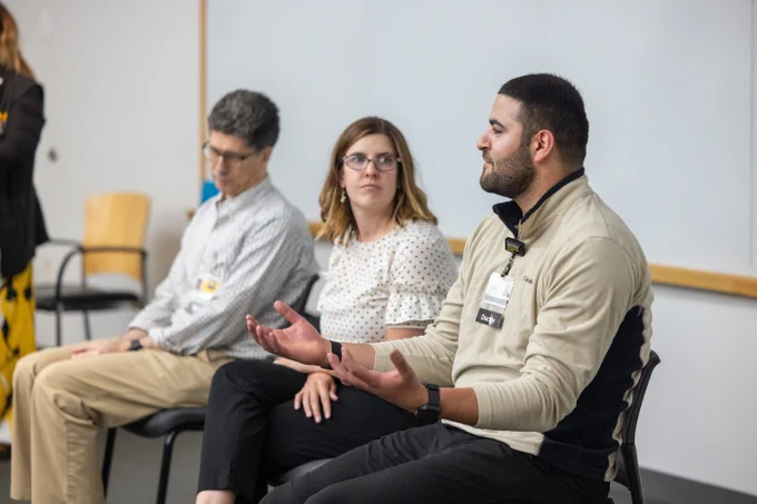 A man in a beige pullover with a name tag gestures while speaking in a panel discussion. Next to him, a woman in glasses and a white polka dot blouse listens attentively, and another man with curly hair and glasses sits with hands clasped. They are seated in a line in a brightly lit room with a whiteboard behind them.