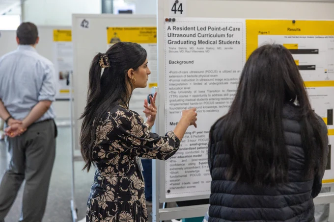 A woman in a floral-patterned dress is presenting a research poster titled "A Resident Led Point-of-Care Ultrasound Curriculum for Graduating Medical Students" to another person. The poster includes text sections labeled "Background" and "Results," featuring graphs and figures. A man in a blue shirt and gray pants stands in the background, looking at another poster.