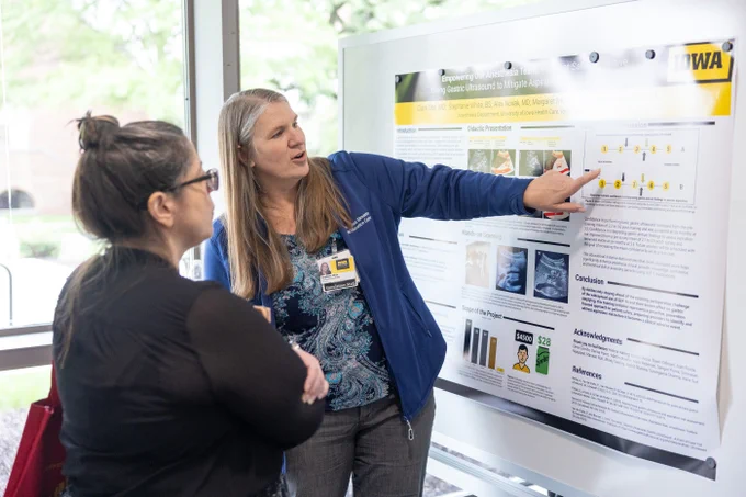 A woman with long hair wearing a blue jacket is presenting a poster to another woman with dark hair and glasses. The poster is titled "Empowering Our Anesthesia Team" and features graphs, images, and text about using gastric ultrasound. They are indoors, with natural light coming through large windows in the background.