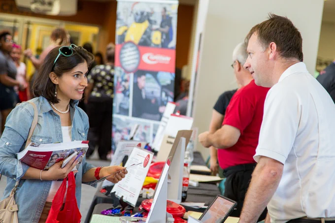 A woman in a denim jacket talks to a man at a busy event booth displaying promotional materials and brochures.