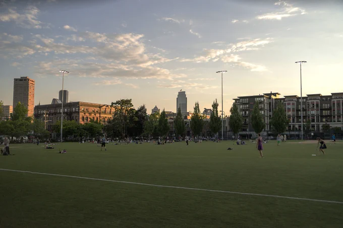 Wide view of a city park with people scattered on the grass and tall buildings in the background under a partly cloudy sky.