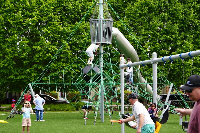 Children playing on a tall rope climbing structure and slide in a green park surrounded by trees on a sunny day.