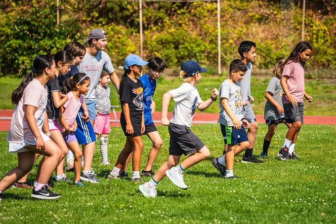 A group of children and teenagers lined up on a grassy field, ready to start a race during a sunny outdoor sports event.