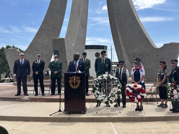 A group of people stands in front of a memorial sculpture at Veterans Memorial Park, some in uniform. A person is speaking at a podium adorned with a symbol. Two wreaths with red, white, and blue flowers are placed nearby. The sky is clear with a few clouds.
