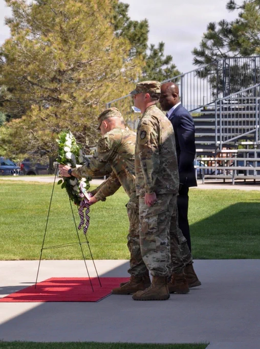 Three people are placing a floral wreath on a stand during a ceremonial event. Two are in camouflage military uniforms, and one is in a suit. They are outdoors, near a grassy field and in front of bleachers.