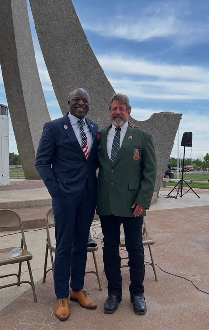 Two people are standing and smiling in front of a large outdoor sculpture. One is wearing a suit and the other a green blazer with a tie. Empty folding chairs and a microphone stand are visible in the background on a paved area. The sky is partly cloudy.