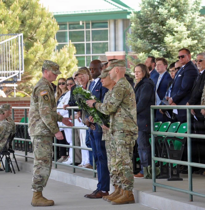 A military ceremony with individuals in uniform participating in a wreath-laying. People in formal attire, including a person in a blue suit, are standing nearby. The event is set in a covered outdoor area with green seating and a backdrop of trees and a building.