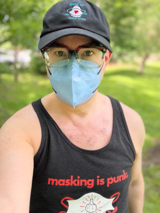Photo of a white man wearing a black cap withdrawing of a white n95 respirator, red, green and clear glasses and a light blue kn95 respirator. He is also wearing a black tank top withdrawing of a white n95 respirator with red straps and drawing of a hand with middle finger flipping up surrounded by red dashes, and above the drawing are the words masking is punk in red text. Green grass background is blurred.