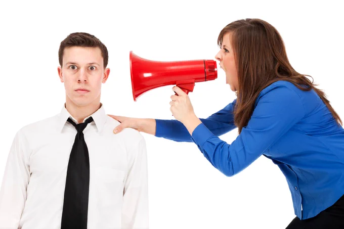 A white man in a shirt and tie sits looking alarmed while a white woman with dark hair and a red megaphone shouts into his ear