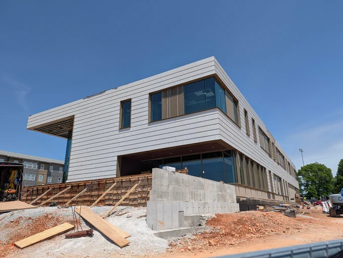 Under construction, the image depicts a modern two-story building with a white exterior. The area surrounding the building is unpaved, with dirt and construction materials visible. The sky is clear and blue.