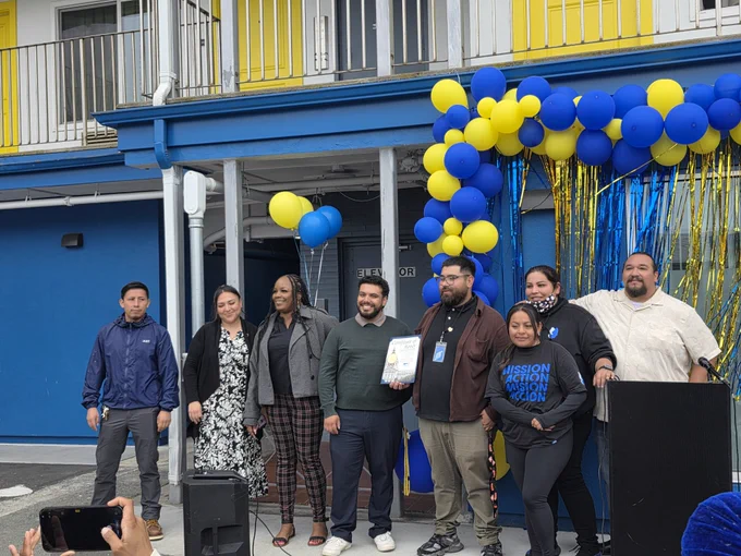 Group photo of community members, partners, and staff standing in front of Casa Colibri during the opening celebration beneath blue and yellow balloon decorations.