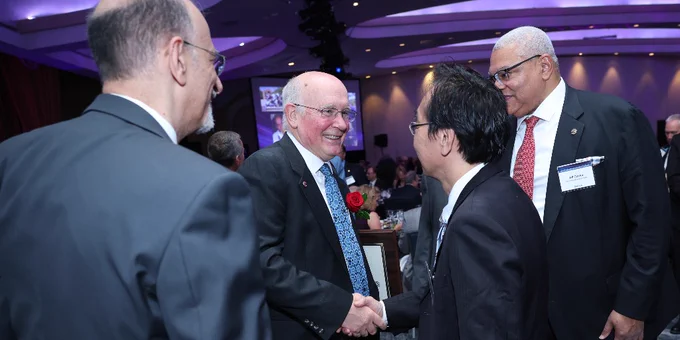 Four individuals are engaged in conversation at a formal event. A person with a rose on their lapel is shaking hands with another. They appear to be in a large venue with dim lighting and a purple-lit ceiling.