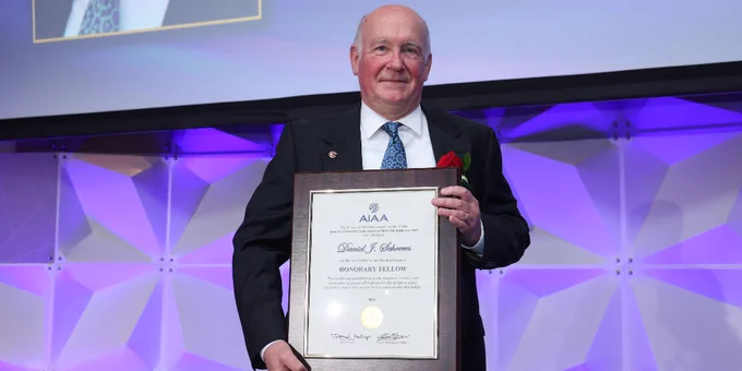 Daniel Scheeres, CU Boulder Distinguished Professor, holding a framed certificate and a red rose lapel pin, standing on a stage with a geometric purple-lit backdrop. The certificate is from AIAA and labeled "Honorary Fellow.