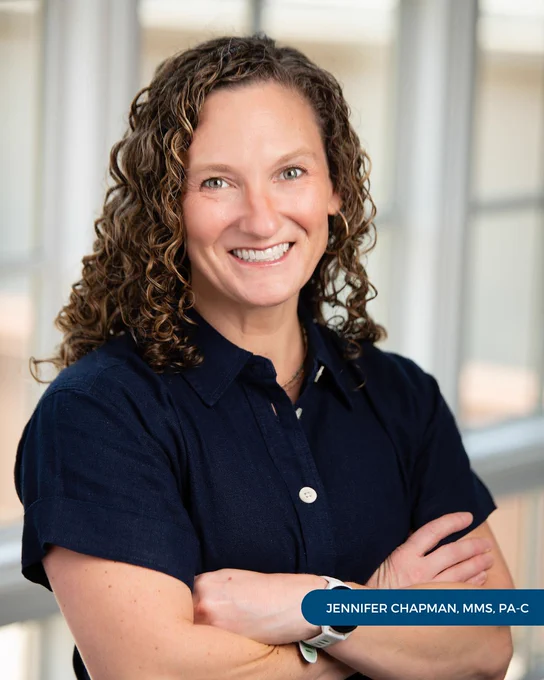 Jennifer Chapman, MMS, PA-C, of Tryon Medical Partners smiles with her arms crossed in a professional portrait posed in front of a paned glass window