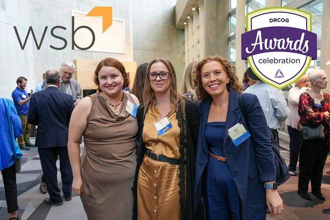 Three women stand and smile together inside a crowded reception area with the logos for WSB and the DRCOG awards celebration overlayed in the upper corners.