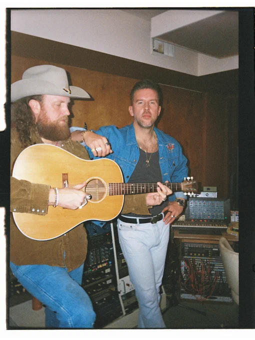 Brother's Osborne are in a studio setting. One is playing an acoustic guitar and wearing a cowboy hat, while the other stands nearby in a denim jacket. The room contains various audio equipment.