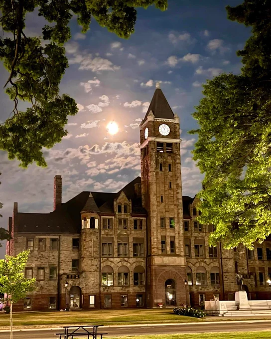 A historic stone building with a tall clock tower is lit by moonlight at night. The sky is partly cloudy, and the ground is surrounded by lush trees.