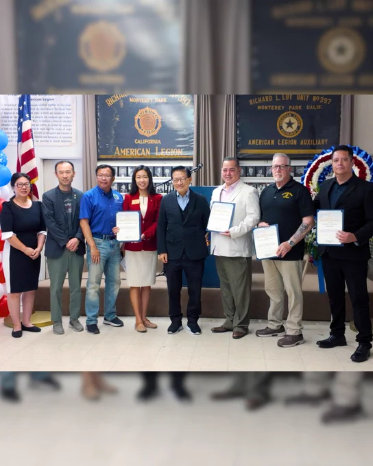 Eight people standing in an American Legion hall; several hold certificates, with US flag and wreath behind.