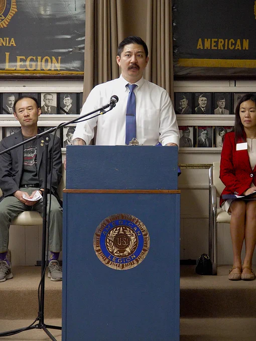 Man in white shirt and blue tie speaking at an American Legion podium with two seated attendees.